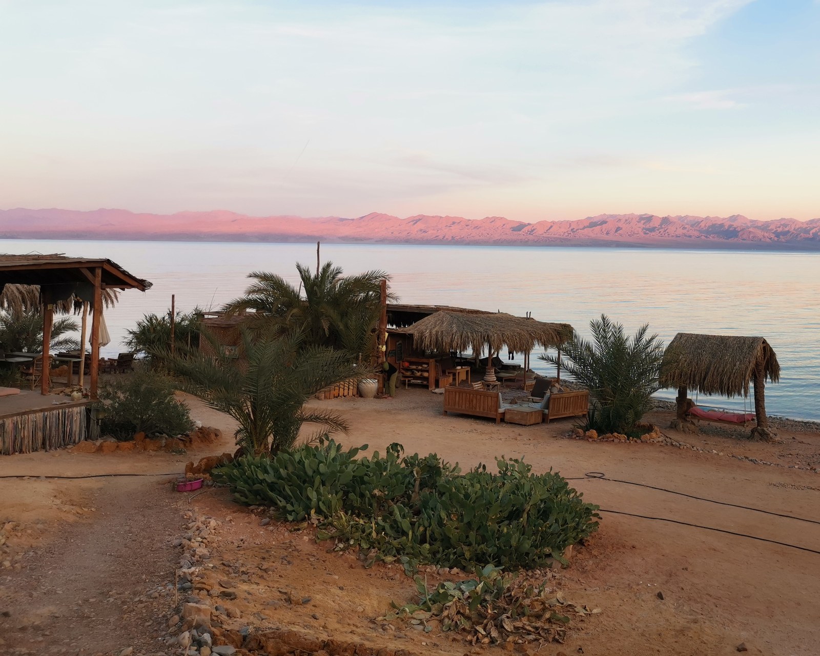 Camp am Meer in der Abenddämmerung, Palmen, Palmdach-Hütten und rosa leuchtende Berge im Hintergrund